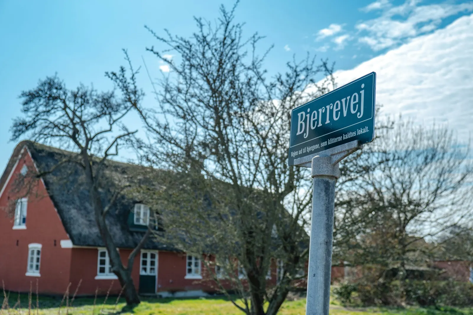 Vejskilt foran et rødt gårdhus med stråtag